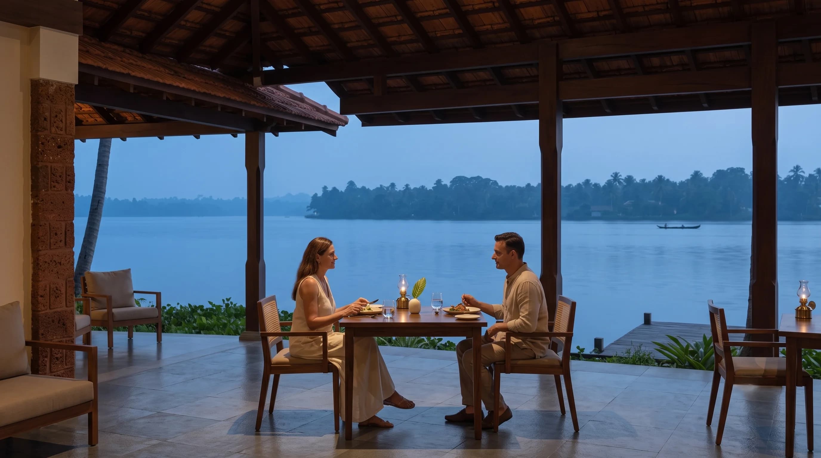 Two guests at a timber table under the tiled dining pavilion at dusk, an oil lamp between them, the lake and far shore beyond.